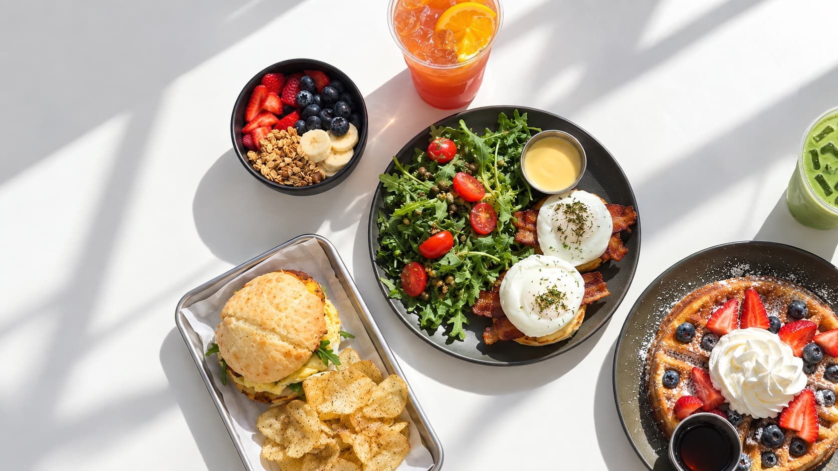 Breakfast at bōm dough in Cambridge, MA: a pão de queijo breakfast sandwich with chips, an açaí bowl with berries and granola, eggs benedict with bacon and arugula salad, a berry mochi waffle, iced matcha and an iced citrus drink.