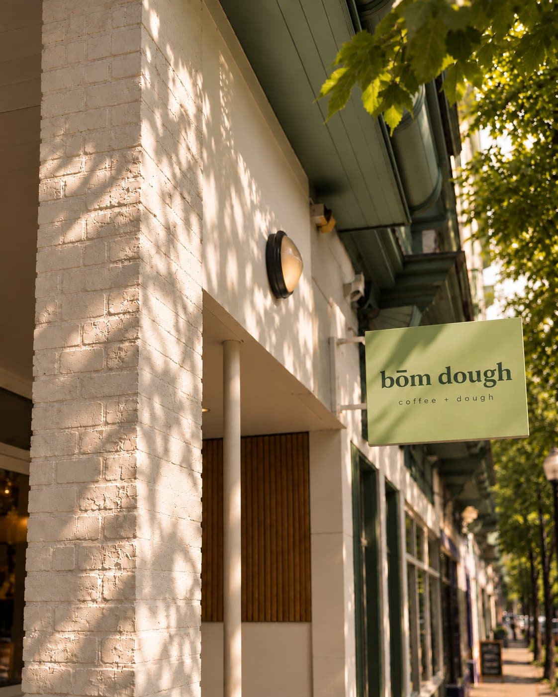 The bōm dough Inman Square café at 1271 Cambridge St, Cambridge, MA — a white-painted brick corner and the green bōm dough · coffee + dough hanging sign.